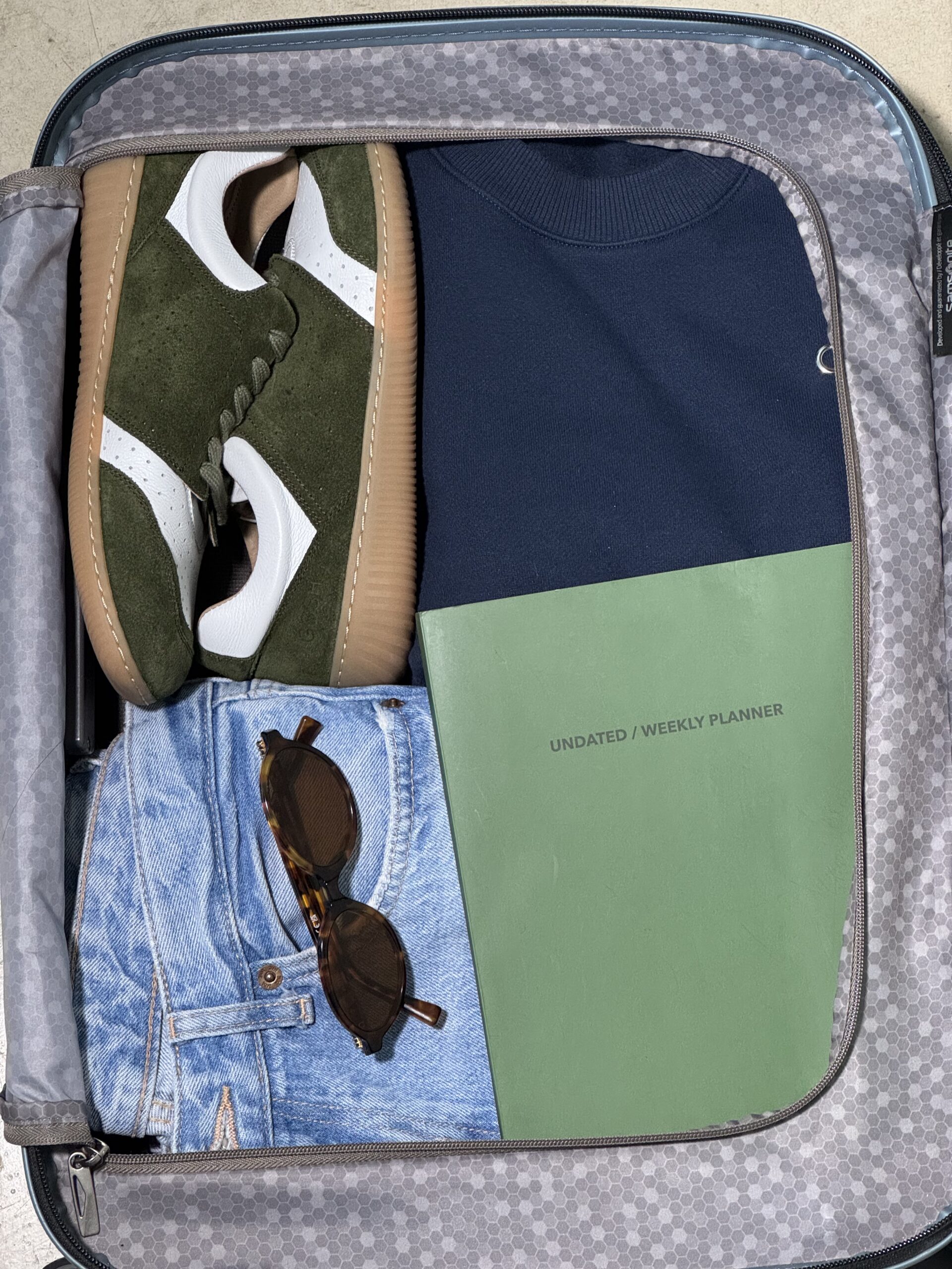 Open travel suitcase neatly packed with green-white GOSH barefoot sneakers, blue hoodie and jeans, sunglasses, and a planner, ready for an organized and stylish trip.