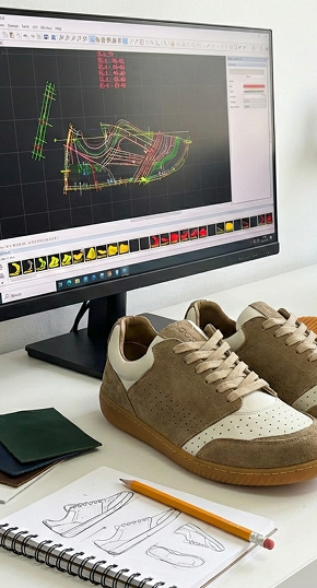 Close view of a professional footwear design workspace featuring beige sneakers on a desk, a sketchbook with design drawings, and a monitor showing CAD modeling software.